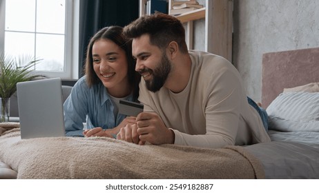 Caucasian happy couple woman and man relax on bed with laptop shopping online use bank credit card pay order buying in internet booking financial service e-commerce app winning success spending money - Powered by Shutterstock