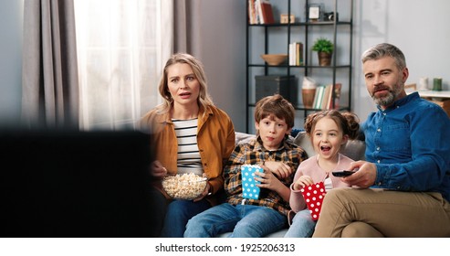 Caucasian happy cute Caucasian family spending time at home watching TV sitting on sofa with popcorn and shocked amazed faces. Parents with small children eating popcorn watching movie or cartoon