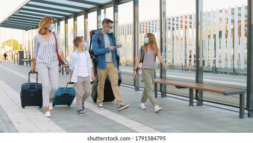 Caucasian happy family with two cute small kids walking at bus stop or train station, carrying suitcases on wheels and talking. Parents with little daughter and son in medical masks travelling.