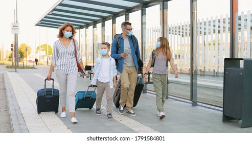 Caucasian happy family with two cute small kids walking at bus stop or train station, carrying suitcases on wheels and talking. Parents with little daughter and son in medical masks travelling.