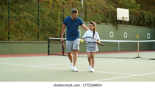 Caucasian happy handsome father with daughter walking away from sport court after playing tennis on sunny hot day. Dad with girl leaving pitch talking, joking and having fun after game.