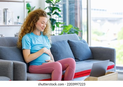 Caucasian Happy Pretty Long Curly Hair Healthy Pregnancy Mother In Casual Outfit Sitting Smiling On Cozy Sofa Touching Rubbing Tummy Belly Watching Online Streaming Via Laptop Computer In Living Room.