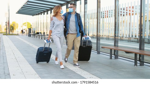 Caucasian happy young married couple in love walking at bus stop and carrying suitcases on wheels. Beautiful woman and handsome man in medical masks hugging and travelling during covid-19 pandemic.