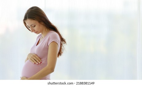 Caucasian Healthy Long Hair Female Prenatal Pregnant Mother Model In Casual Shirt Standing In Front Of Curtain Background In Bedroom Smiling Holding Hands Touching Baby In Big Belly Tummy With Love.