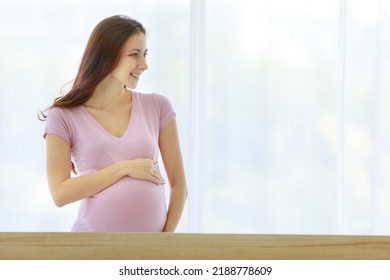 Caucasian Healthy Long Hair Female Prenatal Pregnant Mother Model In Casual Shirt Standing In Front Of Curtain Background In Bedroom Smiling Holding Hands Touching Baby In Big Belly Tummy With Love.