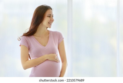 Caucasian Healthy Long Hair Female Prenatal Pregnant Mother Model In Casual Shirt Standing In Front Of Curtain Background In Bedroom Smiling Holding Hands Touching Baby In Big Belly Tummy With Love.