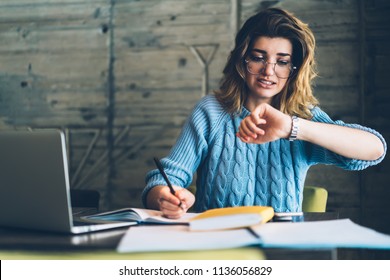 Caucasian hipster girl checking time on wristwatch feeling unhappy and hurrying up on deadline learning and doing homework using literature and laptop at wifi zone, stressed student preparing for exam