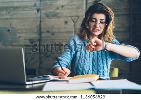 Caucasian hipster girl checking time on wristwatch feeling unhappy and hurrying up on deadline learning and doing homework using literature and laptop at wifi zone, stressed student preparing for exam