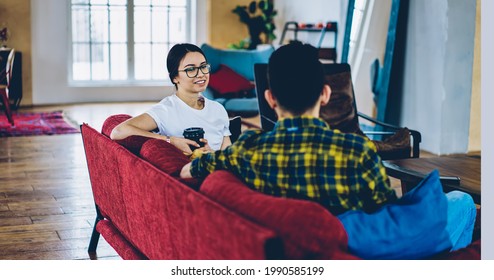Caucasian hipster girl in optical spectacles for provide eyes correction enjoying friendly communication with male friend, young man and woman with coffee resting in living room with red sofa - Shutterstock ID 1990585199