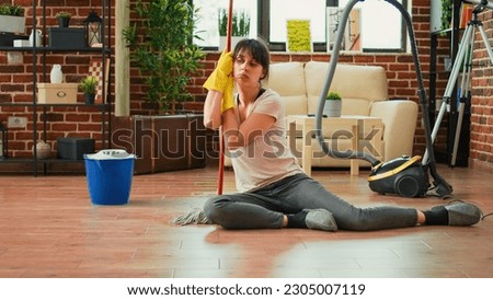 Caucasian housewife feeling sleepy after finishing spring cleaning session, sitting on clean wooden floor with mop and gloves. Tired woman being proud of tidy apartment, household chores.
