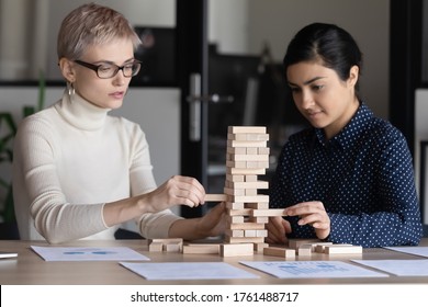 Caucasian Indian Two Businesswomen Sit At Desk In Office Build Tower Of Wooden Blocks Involved In Game Play. Team Building Activity, Teamwork, Business Strategizing, Strategic Thinking Concept