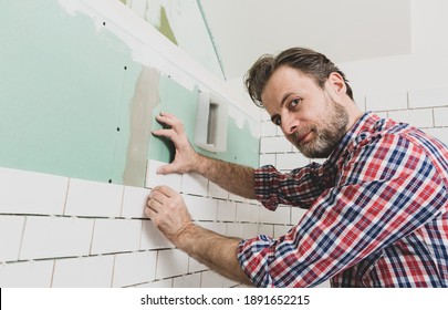 Caucasian interior finishing worker (tiler) laying small white ceramic tiles on the wall. Bathroom renovation works. Handyman in plaid shirt. 