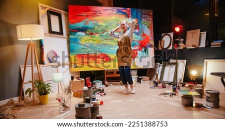 Caucasian joyful young pretty woman painter in apron listening to favorite music in headphones and dancing rhythmically while drawing on big canvas with oil paints in cozy own workshop at night, art