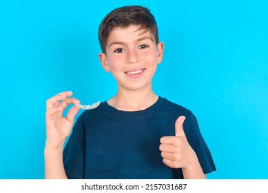 caucasian kid boy wearing blue T-shirt over blue background holding an invisible braces aligner and rising thumb up, recommending this new treatment. Dental healthcare concept.