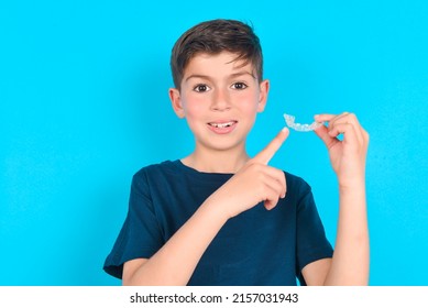 Caucasian Kid Boy Wearing Blue T-shirt Over Blue Background Holding An Invisible Aligner And Pointing At It. Dental Healthcare And Confidence Concept.