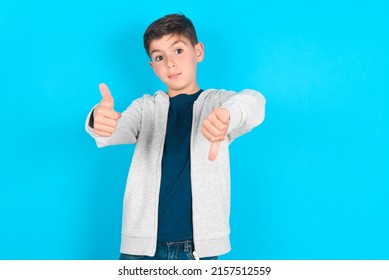 Caucasian Kid Boy Wearing Grey Hoodie Over Blue Background Feeling Unsure Making Good Bad Sign. Displeased And Unimpressed.