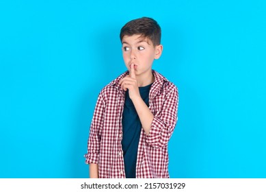 Caucasian Kid Boy Wearing Plaid Shirt Over Blue Background Makes Silence Gesture, Keeps Index Finger To Lips Makes Hush Sign. Asks Not To Share Secret.