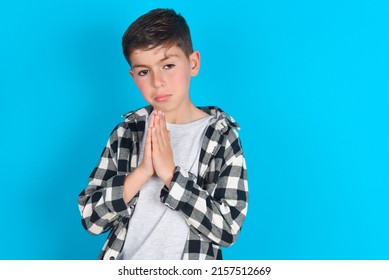 caucasian kid boy wearing plaid shirt over blue background keeps palms pressed together in front of her having regretful look, asking for forgiveness. Forgive me please. - Shutterstock ID 2157512669