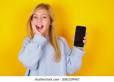 Caucasian Kid Girl Wearing Blue Knitted Sweater Over Yellow Background Hold Hand Modern Technology Use Touch Face Palm Astonished Impressed Scream Wow Omg Unbelievable Unexpected