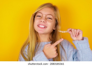 Caucasian Kid Girl Wearing Blue Knitted Sweater Over Yellow Background Holding An Invisible Aligner And Pointing At It. Dental Healthcare And Confidence Concept.