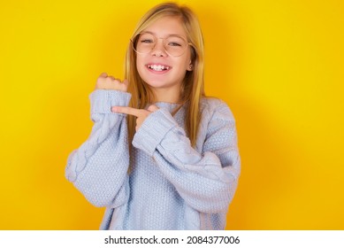 Caucasian Kid Girl Wearing Blue Knitted Sweater Over Yellow Background In Hurry Pointing To Wrist Watch, Impatience, Looking At The Camera With Relaxed Expression