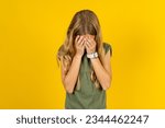 Caucasian kid girl wearing green T-shirt over yellow background covering her face with her hands, being devastated and crying. Sad concept