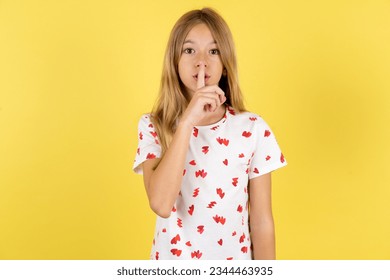 Caucasian kid girl wearing polka dot shirt over yellow background makes silence gesture, keeps finger over lips. Silence and secret concept. - Shutterstock ID 2344463935