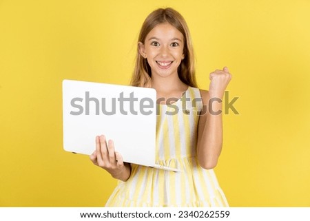 Caucasian kid girl wearing yellow dress over yellow background hold computer open mouth rise fist