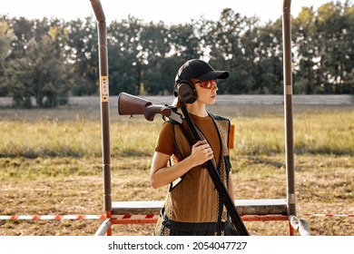 caucasian lady with rifle weapon in outdoor academy shooting range, field in the background. Young beautiful woman in cap, protective headset and spectacles posing, ready to train, looking at side