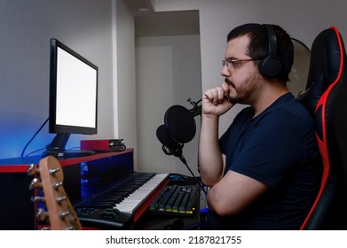 Caucasian Latino Music Engineer Man, With Headphones And Glasses, Is With One Hand On His Chin And His Arms Crossed Serious And Thoughtful, Sitting Watching The Monitor In His Home Studio