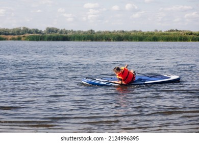 Caucasian little female kid sitting on sup board on small lake trying to stand up on feet in life jacket. Active holidays full of adventures. Inculcation of love for sports from childhood. - Shutterstock ID 2124996395