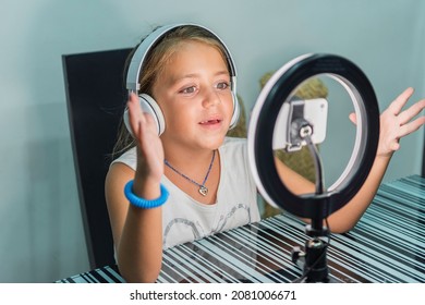 Caucasian Little Girl Making A Video Tutorial With Mobile Phone, Headphones And Light Ring With Tripod At Home.