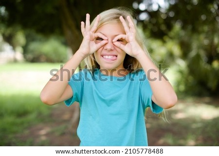 caucasian little girl wearing blue t-shirt standing outdoors doing ok gesture like binoculars sticking tongue out, eyes looking through fingers. Crazy expression.