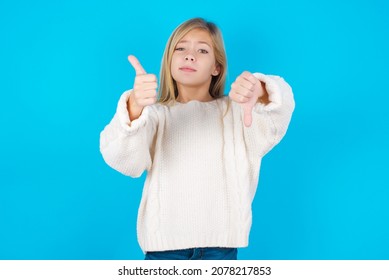 Caucasian Little Kid Girl Wearing White Knitted Sweater Over Blue Background Feeling Unsure Making Good Bad Sign. Displeased And Unimpressed.