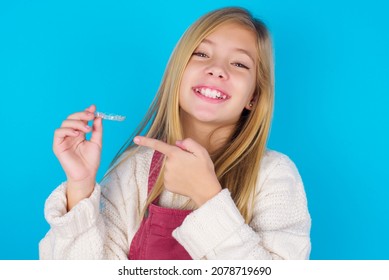 Caucasian Little Kid Girl Wearing Jumpsuit Over Blue Background Holding An Invisible Aligner And Pointing At It. Dental Healthcare And Confidence Concept.