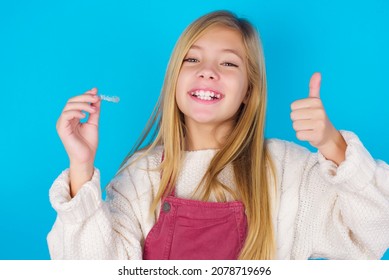 Caucasian Little Kid Girl Wearing Jumpsuit Over Blue Background Holding An Invisible Braces Aligner And Rising Thumb Up, Recommending This New Treatment. Dental Healthcare Concept.