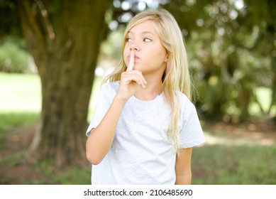 Caucasian Little Kid Girl Wearing T-shirt Standing Outdoors Silence Gesture Keeps Index Finger To Lips Makes Hush Sign. Asks Not To Share Secret