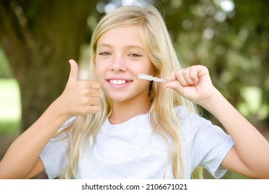 Caucasian Little Kid Girl Wearing T-shirt Standing Outdoors Holding An Invisible Braces Aligner And Rising Thumb Up, Recommending This New Treatment. Dental Healthcare Concept.