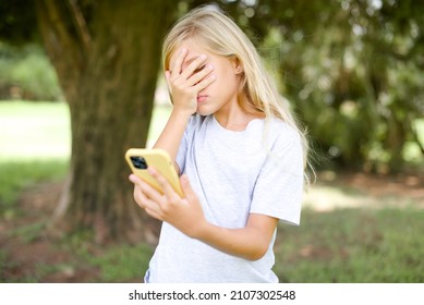 Caucasian Little Kid Girl Wearing T-shirt Standing Outdoors Looking At Smart Phone Feeling Sad Holding Hand On Face.