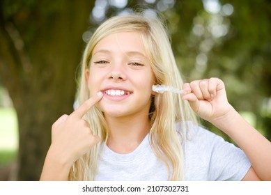 Caucasian Little Kid Girl Wearing T-shirt Standing Outdoors Holding An Invisible Aligner And Pointing To Her Perfect Straight Teeth. Dental Healthcare And Confidence Concept.