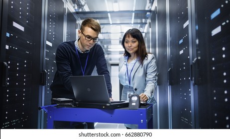 Caucasian Male And Asian Female IT Technicians Working With Computer Crash Cart In Big Data Center Full Of Rack Servers.