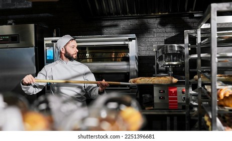 Caucasian male baker in apron and hat taking out just-baked baguettes from oven and putting on shelf in kitchen of bakehouse. Man working in bakery and baking fresh bread. Cooking profession. - Shutterstock ID 2251396681