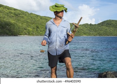 Caucasian male castaway wearing dirty wet clothes and sea grapes hat holds wooden fishing tool while gazing toward the sun on tropical island - Shutterstock ID 496403590