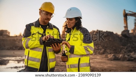 Caucasian Male Civil Engineer Talking To Hispanic Female Inspector And Using Tablet Computer On Construction Site of a New Building. Real Estate Developers Discussing Business, Excavators Working.