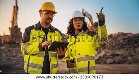 Caucasian Male Civil Engineer Talking To Hispanic Female Inspector And Using Tablet Computer On Construction Site of New Building. Real Estate Developers Discussing Business, Excavators Working.