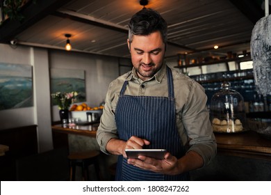 Caucasian Male coffee shop owner smiling at digital tablet while standing in coffee shop, happy with current sales, successful and happy to be open