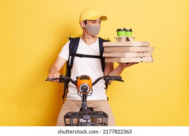 Caucasian male courier on bike overloaded with carton pizza boxes and takeaway coffee cups, dressed in white t shirt, cap and protective mask, working during quarantine isolated over yellow background