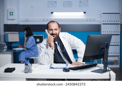 Caucasian male doctor seated at the office desk talking on the telephone with other healthcare specialists. Man wearing a lab coat is using a landline phone to speak with a hospital receptionist. - Powered by Shutterstock