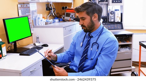 Caucasian male doctor wearing blue scrubs reviewing document. He has dark hair, a beard, and is sitting by a desk with a green screen monitor - Powered by Shutterstock