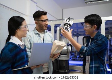 Caucasian Male Engineer Standing Hold A Robot Controller Next To Asian Female Apprentice Holding A Laptop, Demonstrating The Work Of Robotic Machine To Young Asian Male CEO Who Smile Touching A Robot.